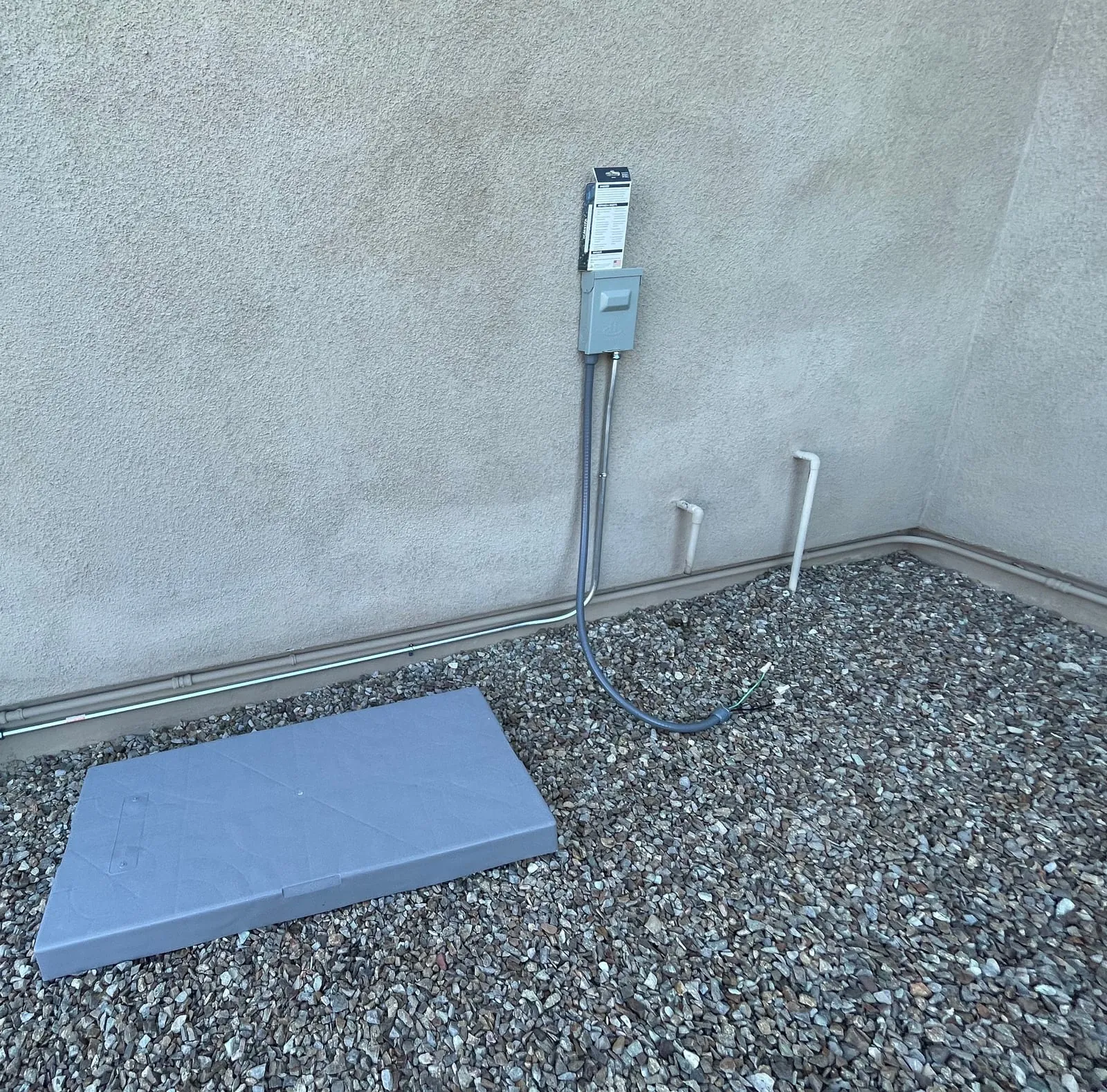 A new equipment pad on the gravel beside the stucco wall, with the electrical run and disconnect in place.