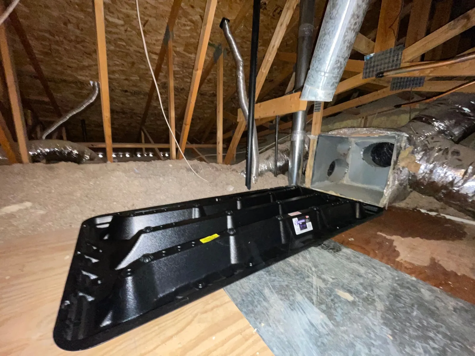 A black emergency drain pan set on the attic floor beside the duct plenum.